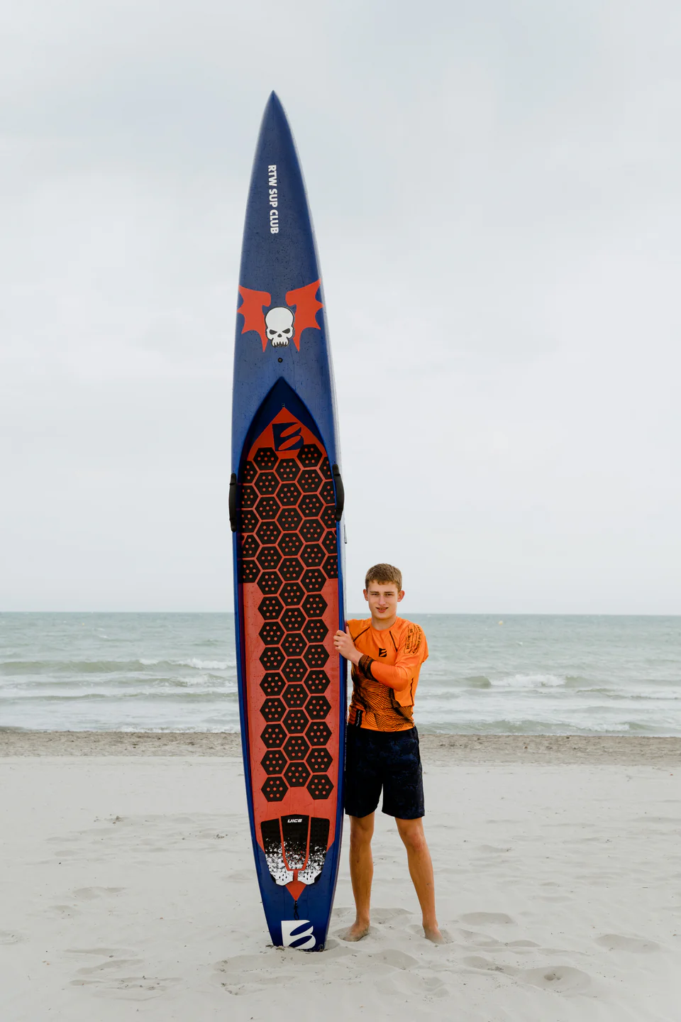 Chłopak stoi na plaży z niebieską sztywną deską SUP z karbonu marki Bass Polska